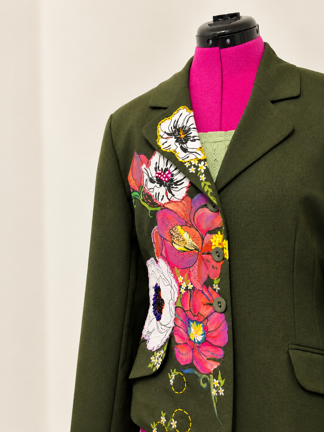 An olive green wool blazer with colorful floral embroidery, displayed on a black dress form mannequin against a plain off-white background.