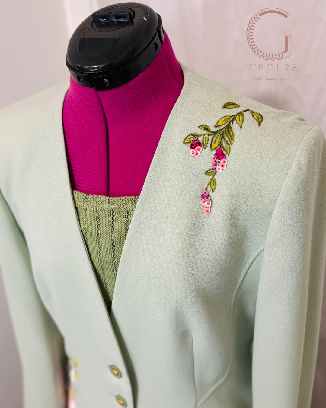 A pale mint green tailored jacket displayed on a magenta dress form, featuring embroidered floral details on the left shoulder with green leaves and pink flowers. A green knit top is layered underneath, set against a light pink backdrop with a Groera watermark.
