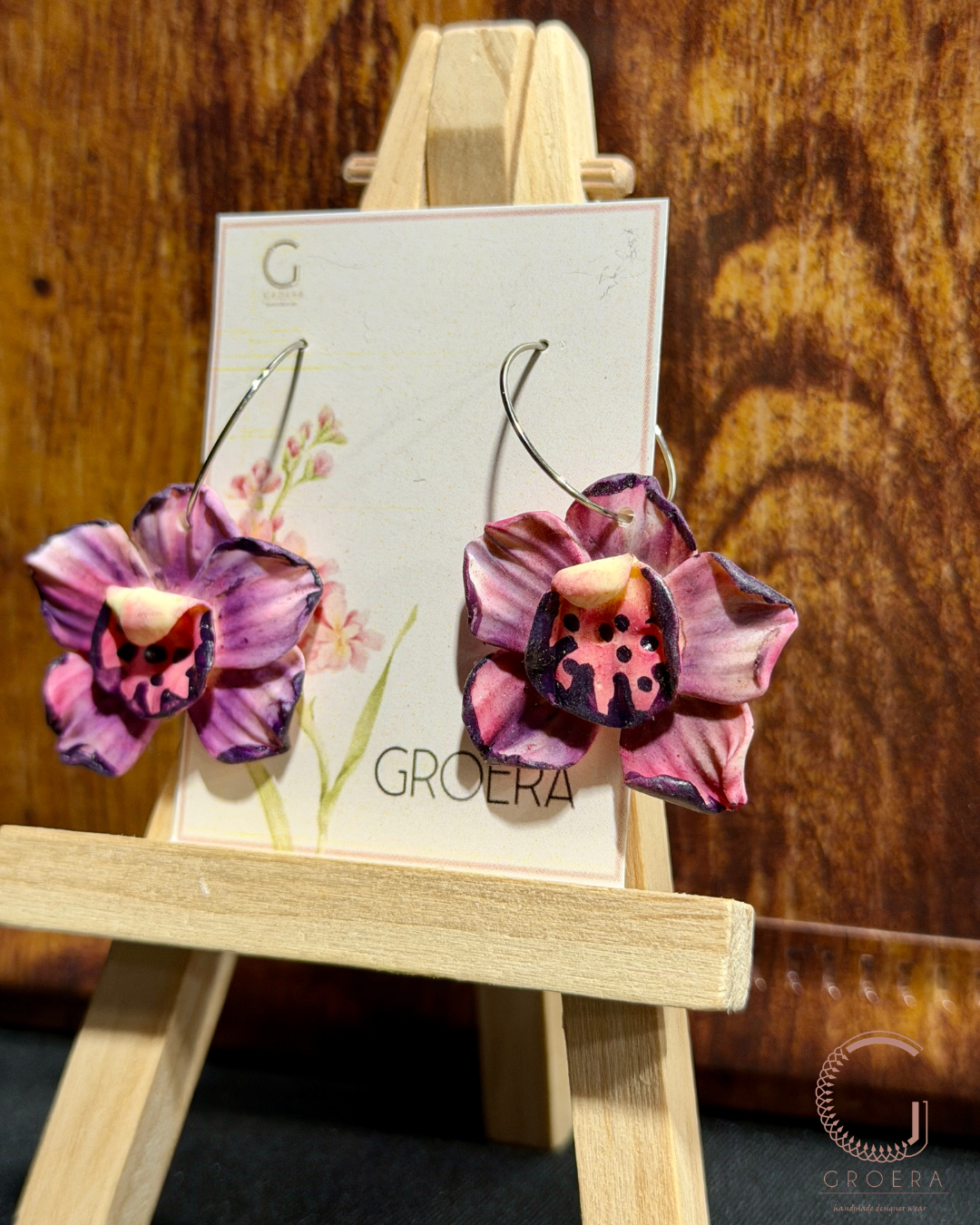 Purple flower-shaped earrings on a Groera branded card with a wooden stand against a wooden background.