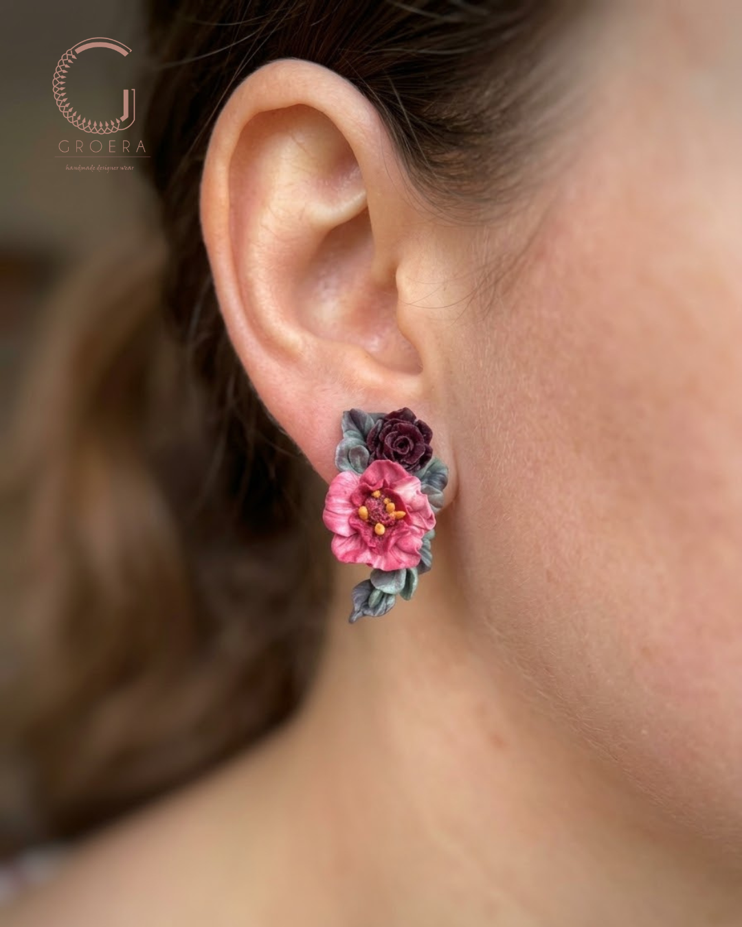 A close-up of a person's ear wearing a floral stud earring with a pink flower, a dark burgundy rose, and gray-green leaves. The Groera brand logo is visible in the top left corner.