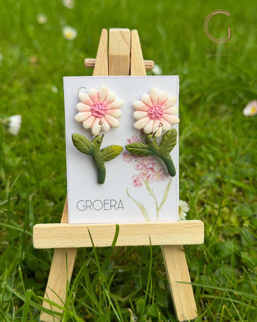Flower-shaped earrings on a card with 'Groera' branding, displayed on a small wooden easel against a grassy background.