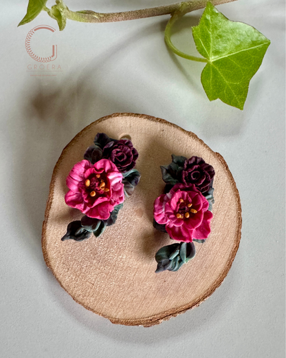 A pair of handmade floral earrings featuring vibrant magenta-pink flowers with dark purple roses and green leaves, displayed on a round wooden slice coaster with a green ivy leaf in the background.