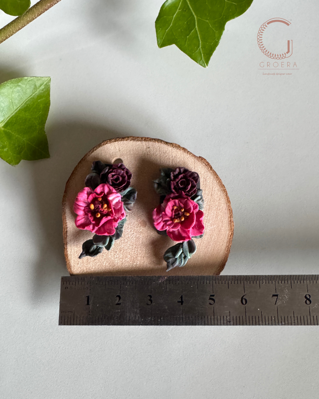 A pair of handmade polymer clay earrings shaped like pink and dark purple flowers with green leaves, displayed on a small round wood slice above a metal ruler for scale, with green ivy leaves in the background.