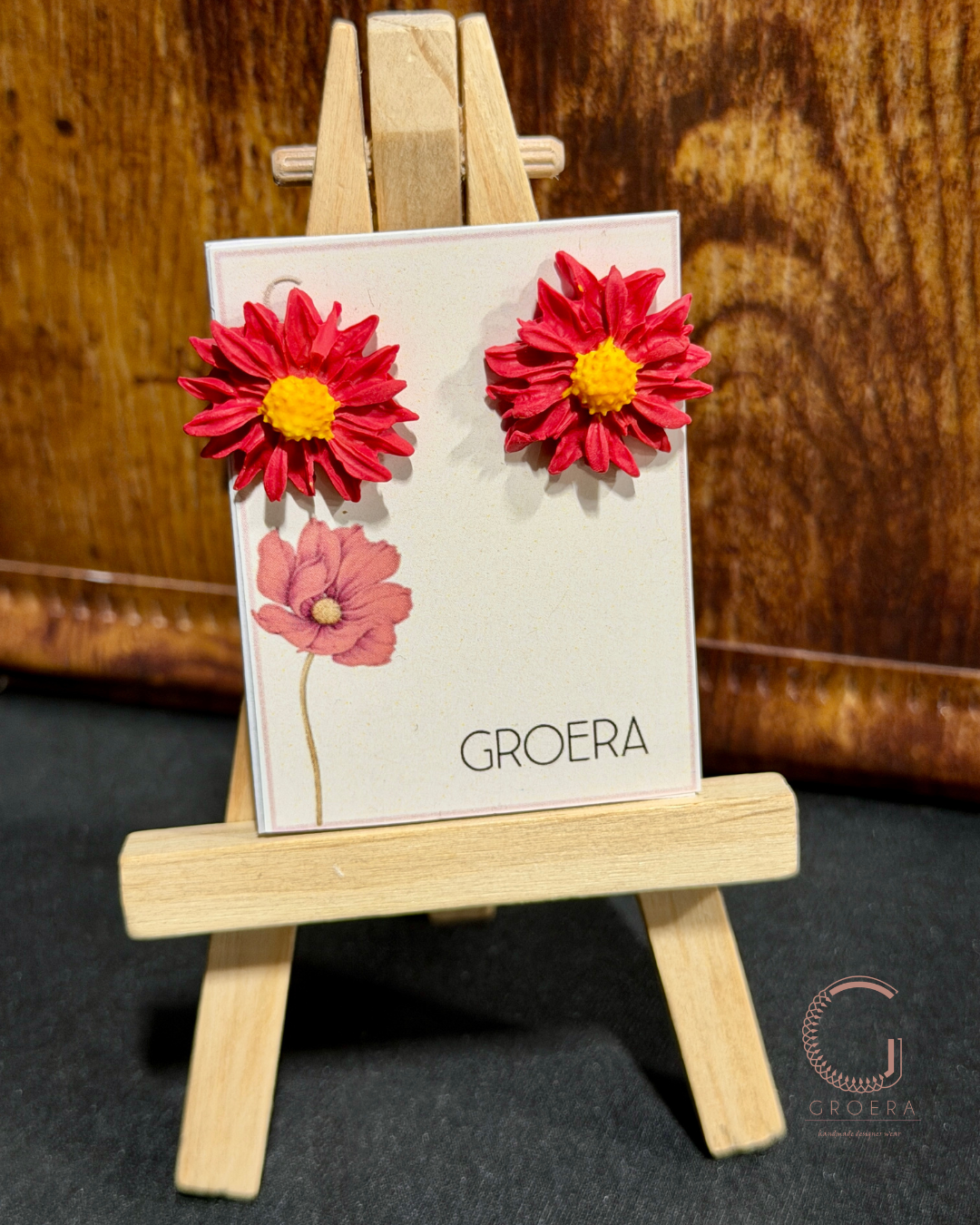 Flower-shaped earrings on a card with 'GROERA' branding, displayed on a small wooden easel.