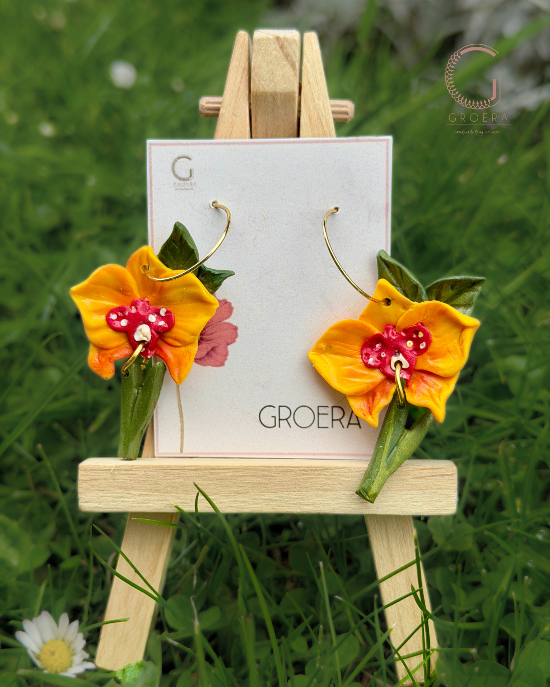 Floral earrings on a small wooden easel with a 'Groera' card against a grassy background.