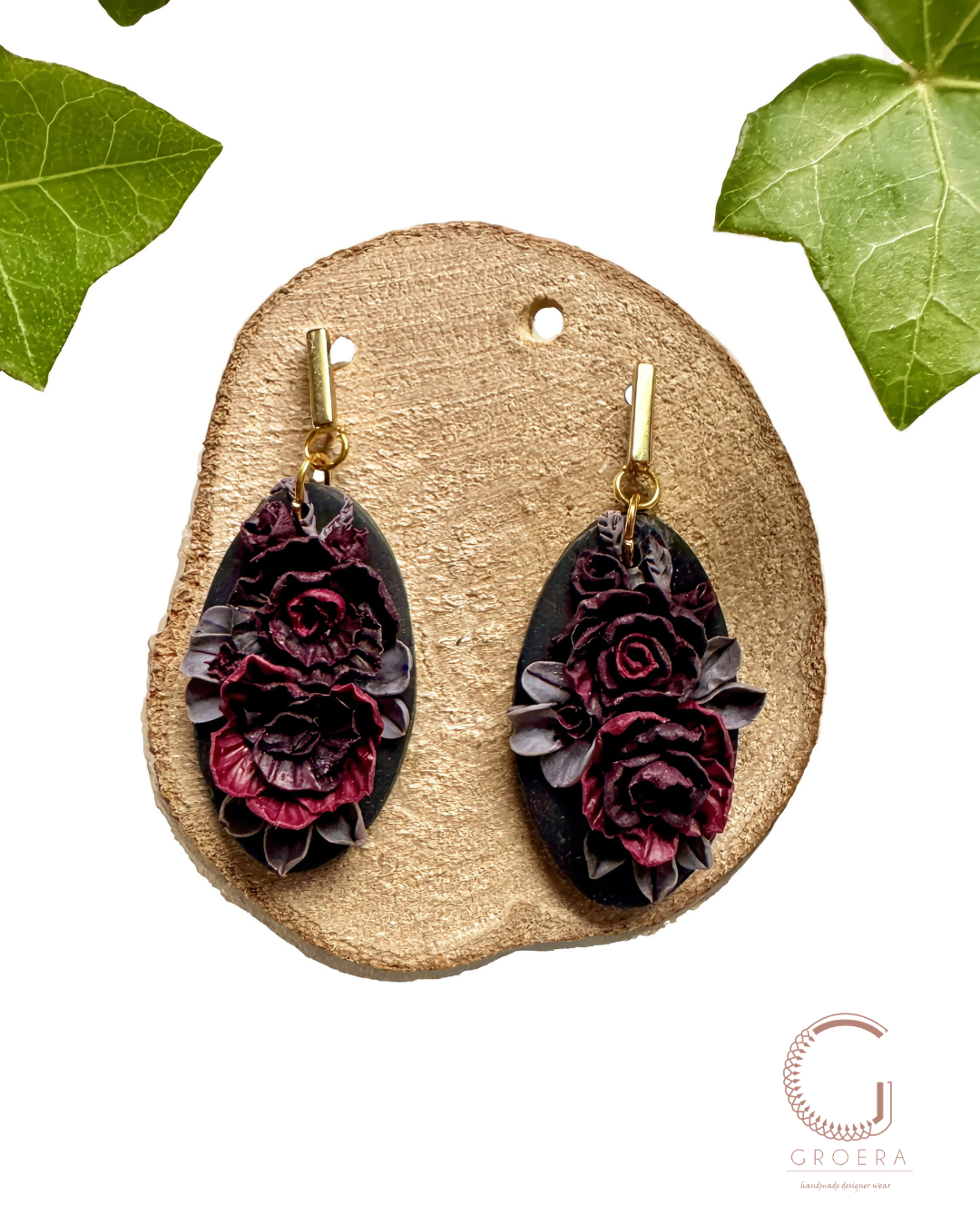 A pair of dangling earrings displayed on a round wood slice against a white background, with green leaves in the corners. The earrings have dark oval pendants featuring layered dark red and purple roses on a black base, with gold-tone hooks.