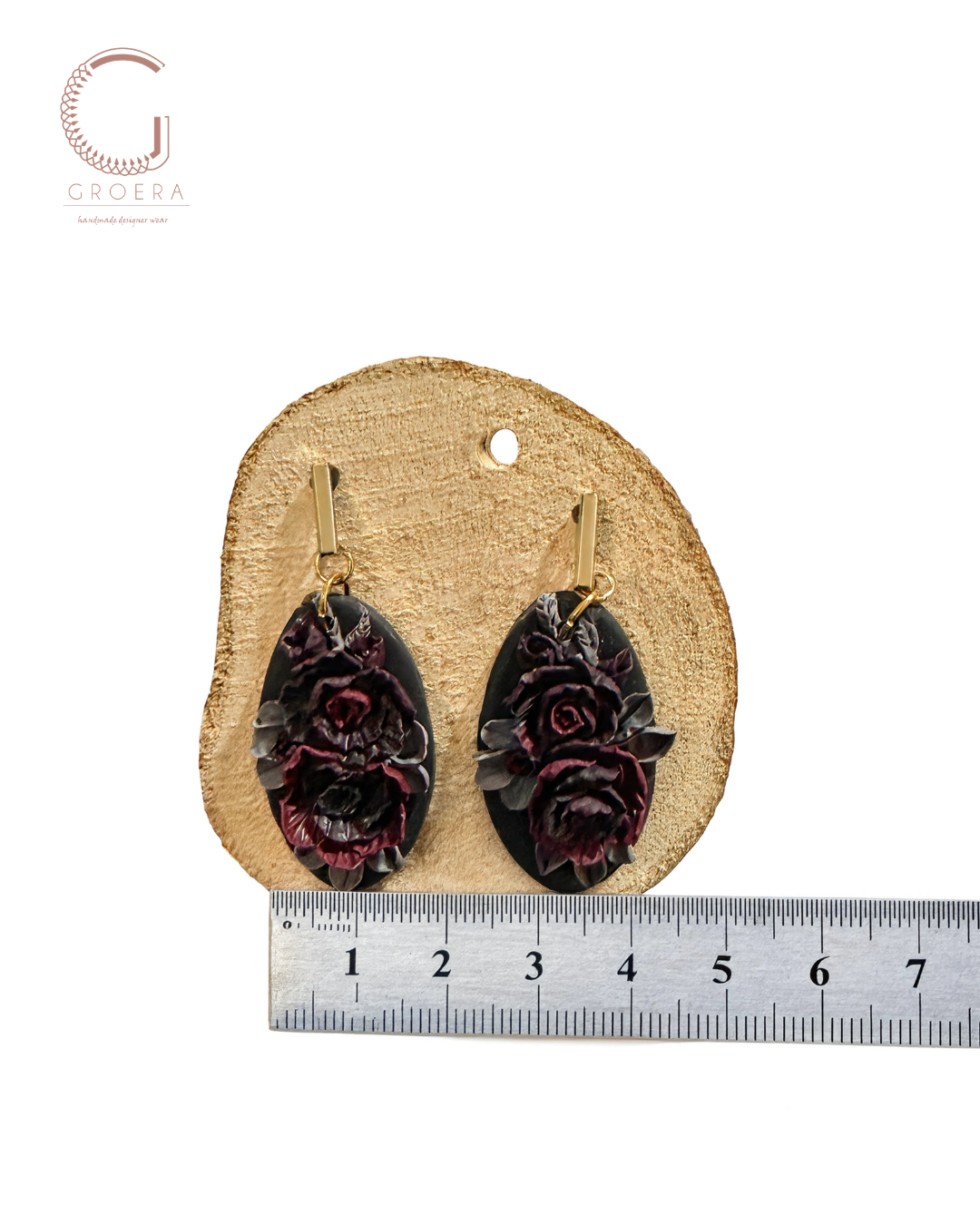 A pair of dangling earrings displayed on a round wooden slice with a metal ruler below for scale. The earrings have dark oval stone bases with raised dark red and burgundy floral carvings and gold-tone hooks.