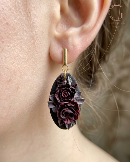 A close-up of a person's ear wearing a dangling earring. The earring has a gold-toned post and a dark oval pendant featuring sculpted dark purple and black roses with muted gray-green leaves.