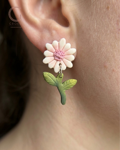 Floral earrings with a pink flower and green leaves worn on an ear.