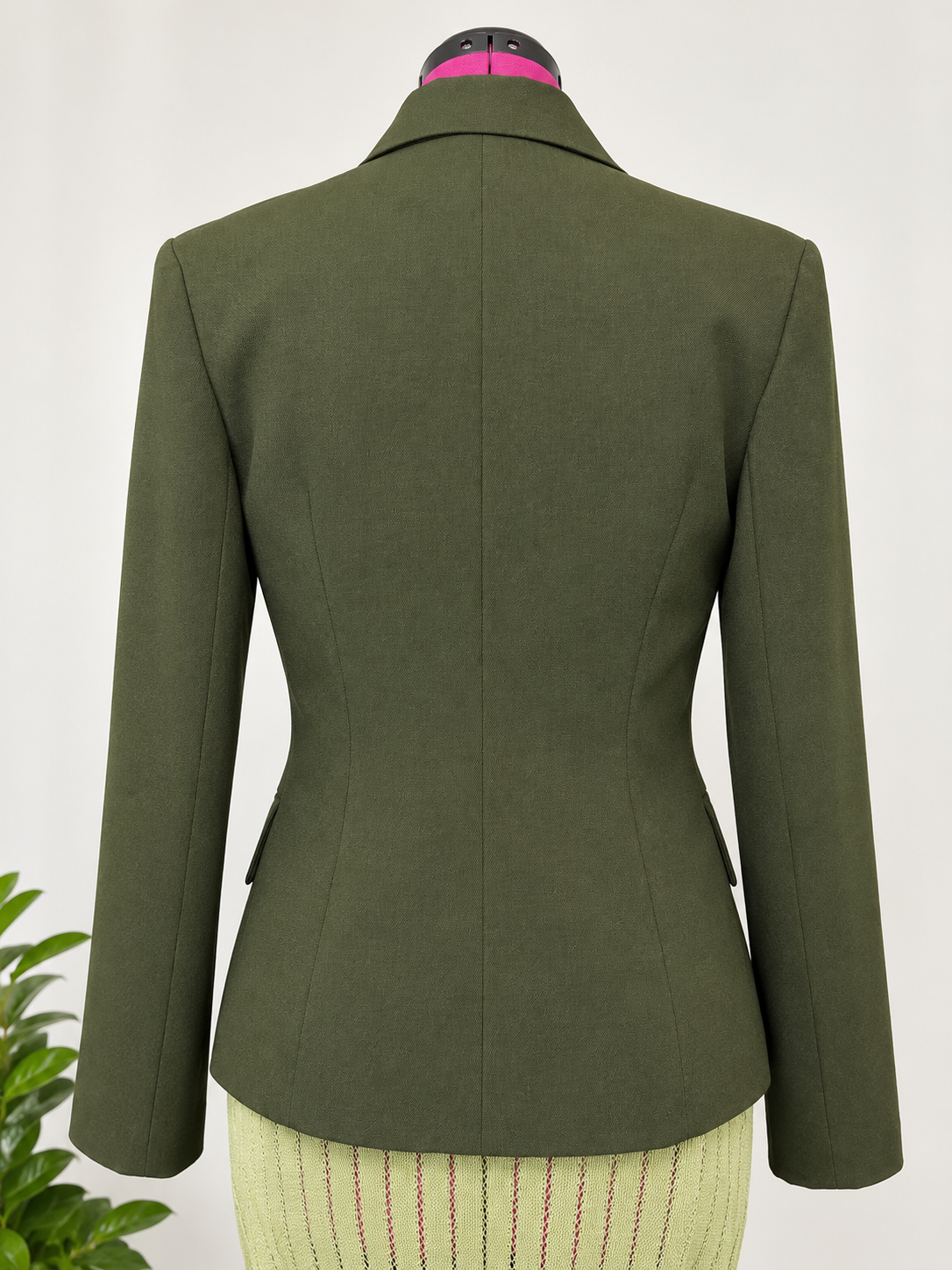 An olive green tailored blazer shown from the back on a dress form mannequin, with a cream striped skirt partially visible underneath.