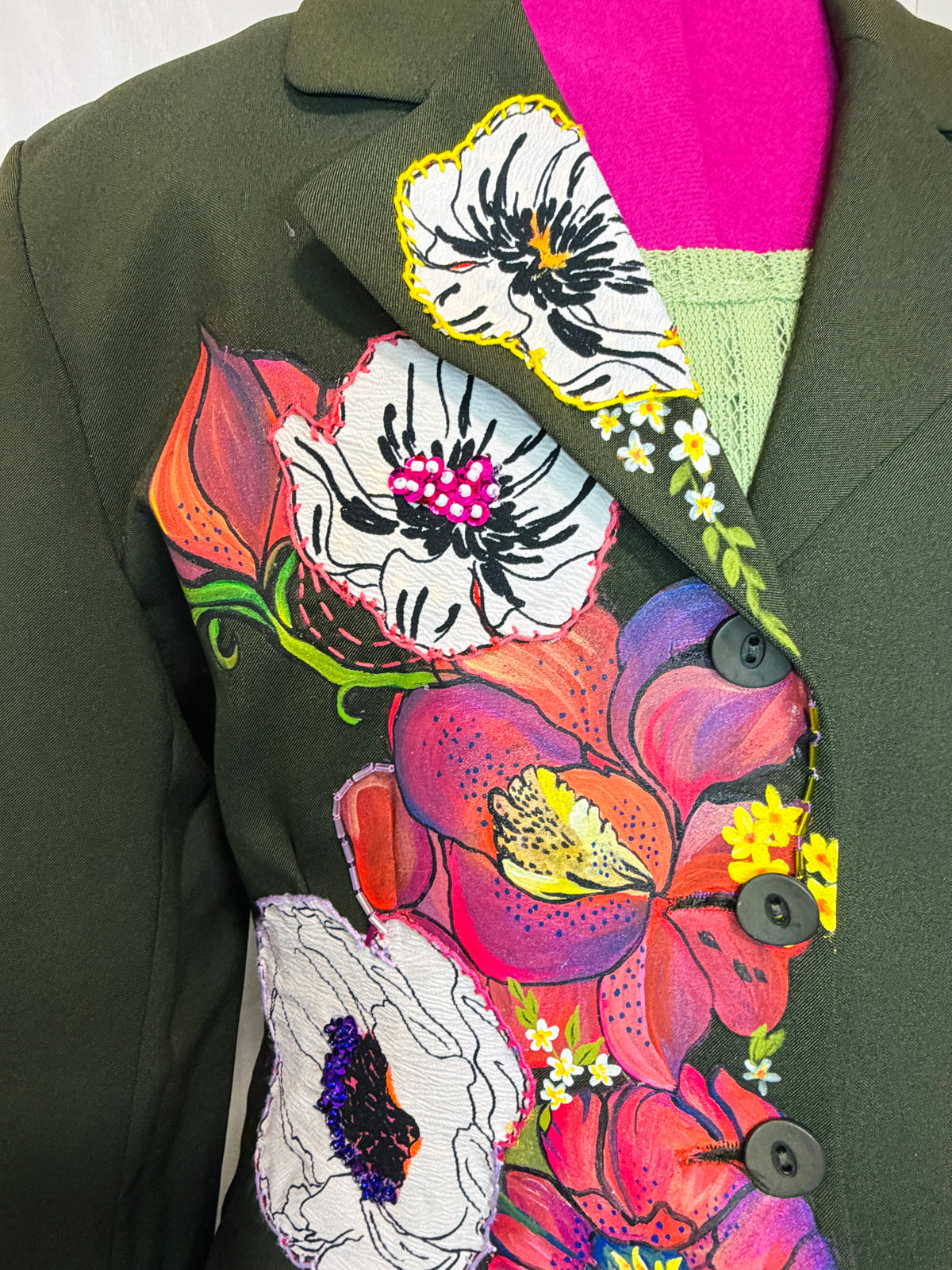 An olive green blazer jacket featuring a large, colorful floral print on the front, layered over a pink and light green knit top. The blazer has dark buttons and is shown on a mannequin torso.