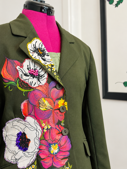 An olive green tailored jacket displayed on a bright pink mannequin torso, featuring large colorful floral appliqués in pink, red, white, and yellow across the front.