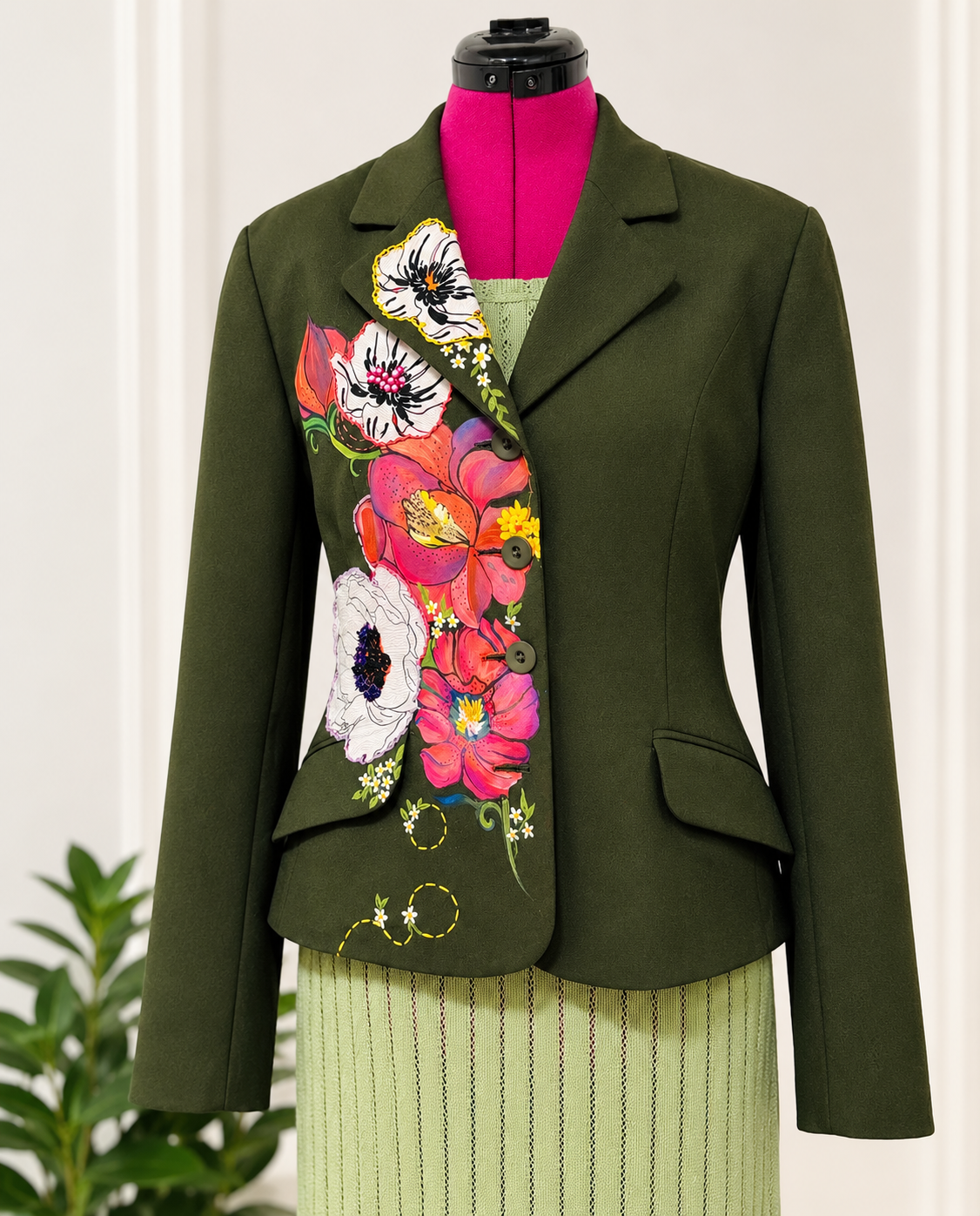 An olive green tailored blazer displayed on a mannequin torso, featuring a large, colorful floral print on the front panel with pink, red, white, and yellow flowers. The blazer is styled with a bright pink top and a light green pleated skirt underneath.