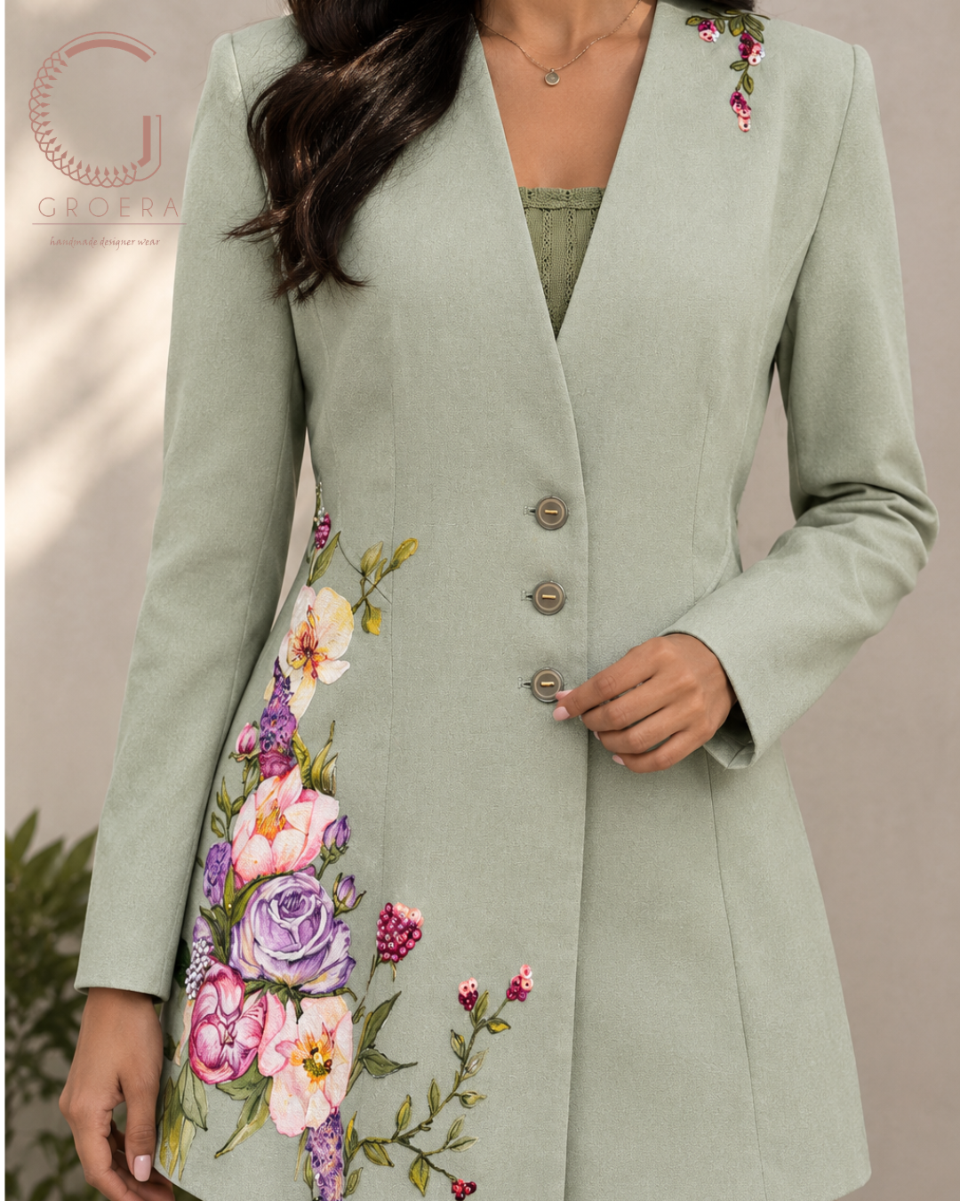 A woman wearing a sage green longline blazer with colorful floral embroidery on the front and shoulder, paired with a green top. The image is cropped from the chest down, showing the fit and details of the garment.