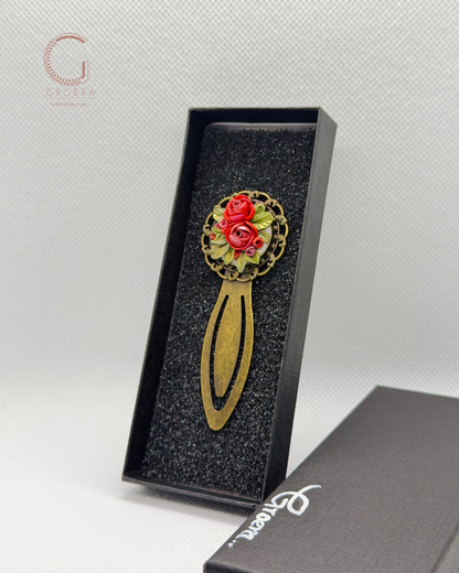 A decorative hair claw clip with a gold-tone metal base and a circular floral head featuring red roses and green leaves, displayed in an open black presentation box.