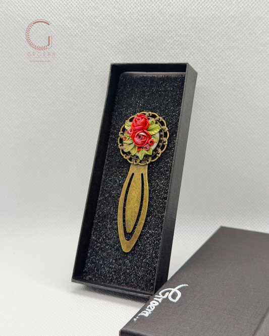 A decorative hair claw clip with a gold-tone metal base and a circular floral head featuring red roses and green leaves, displayed in an open black presentation box.