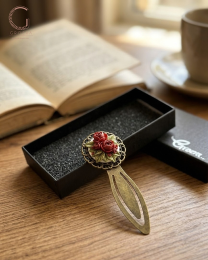 An antique-style metal bookmark with a red rose and green leaves design, resting in an open black presentation box on a wooden table with an open book and a cup of coffee in the background.