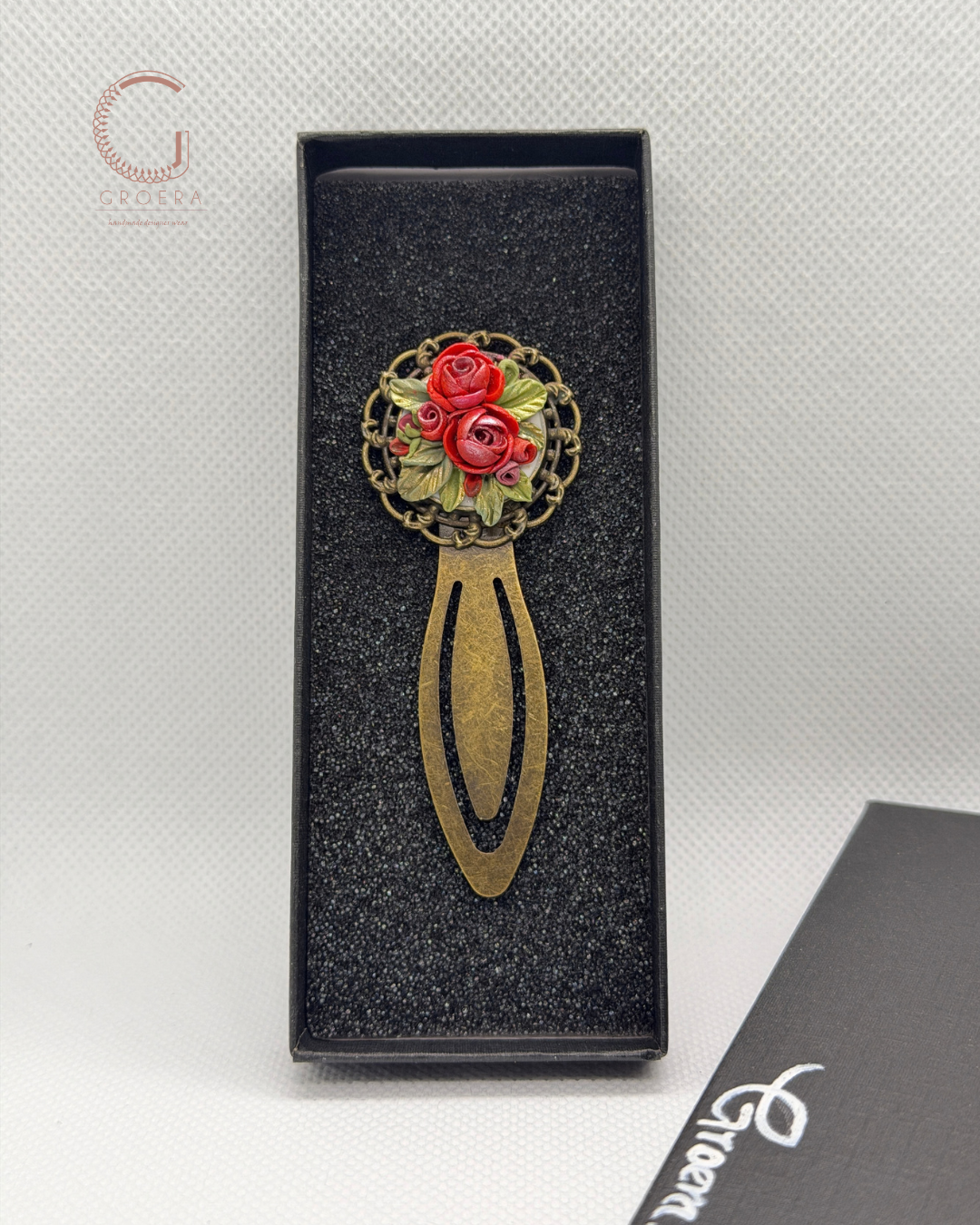 A decorative metal hair pin with a circular floral design featuring red roses and green leaves, set in an ornate gold-tone frame and attached to a long, curved gold-tone handle. The pin is displayed inside an open black presentation box on a textured light gray background.
