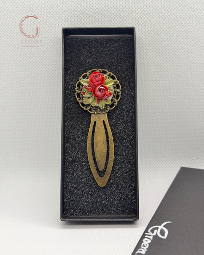 A decorative metal hair pin with a circular floral design featuring red roses and green leaves, set in an ornate gold-tone frame and attached to a long, curved gold-tone handle. The pin is displayed inside an open black presentation box on a textured light gray background.