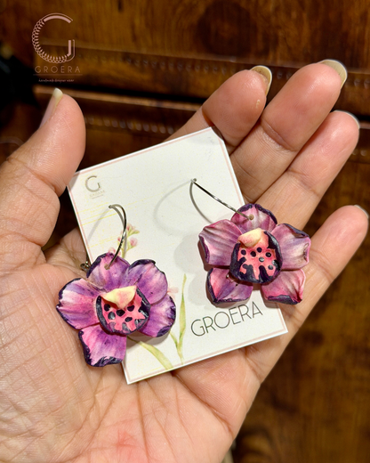Purple flower-shaped earrings held by a hand with a 'Groera' card in the background.