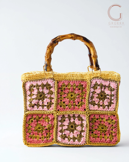 A crocheted handbag with a rectangular shape, featuring six colorful granny square panels in pink, yellow, and olive green, and a curved natural bamboo handle, shown against a light gray background.