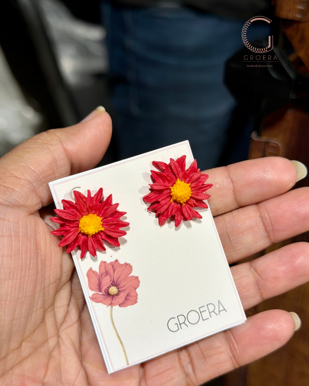 Red flower-shaped earrings on a card held by a hand, with 'GROERA' branding.