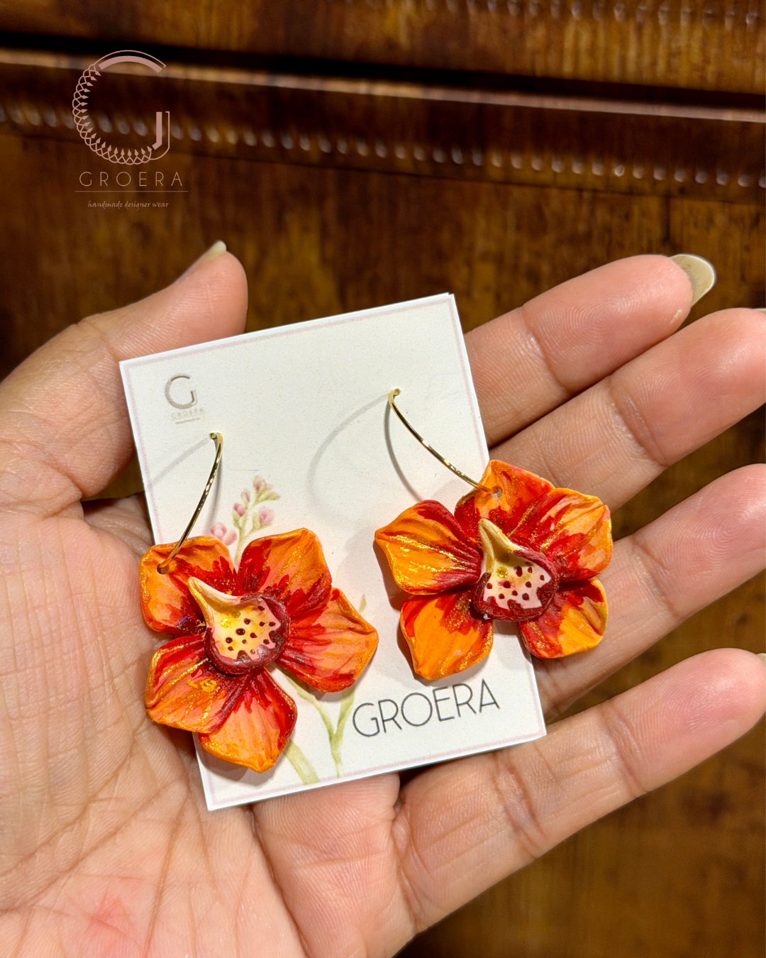Orange flower earrings held by a hand with a Groera card in the background.