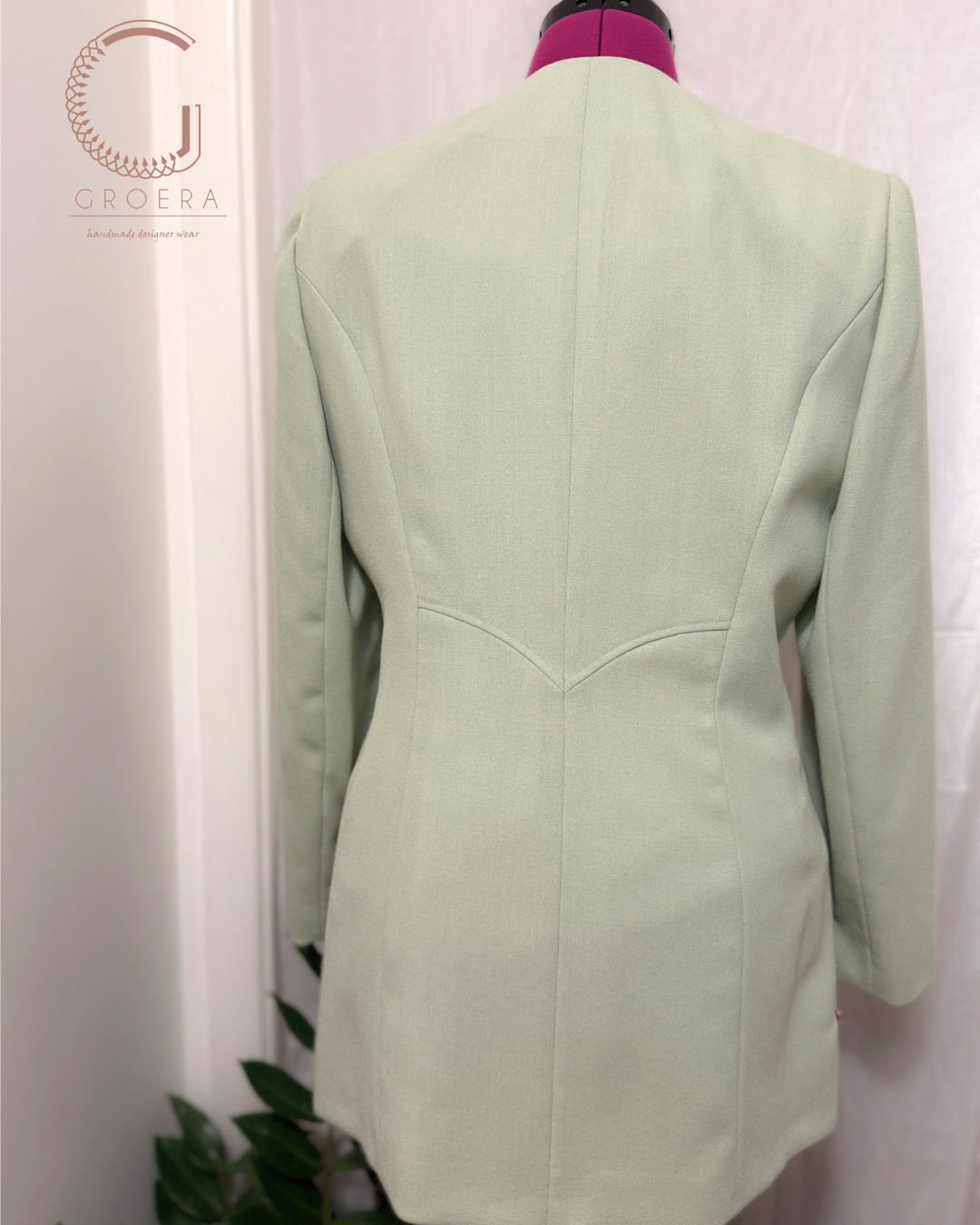 A sage green tailored blazer shown from the back on a mannequin torso with a magenta neck cover, photographed against a white curtain backdrop.