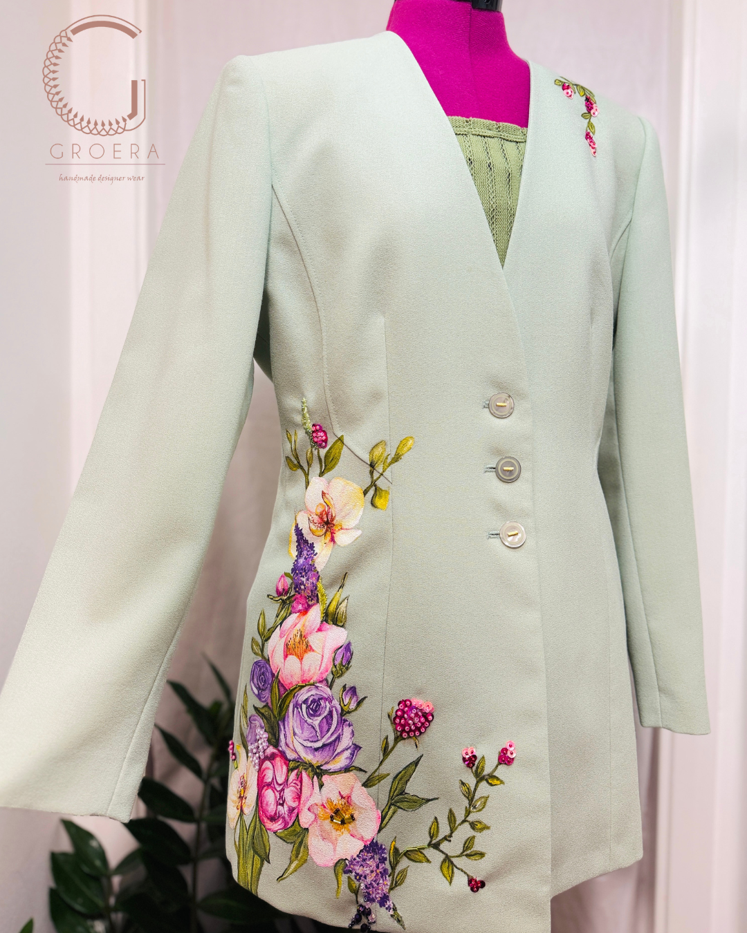A pale mint green tailored blazer with colorful floral embroidery on the front, displayed on a pink mannequin torso. The blazer features three front buttons and is shown against a light curtain backdrop with a Groera logo watermark in the upper left.