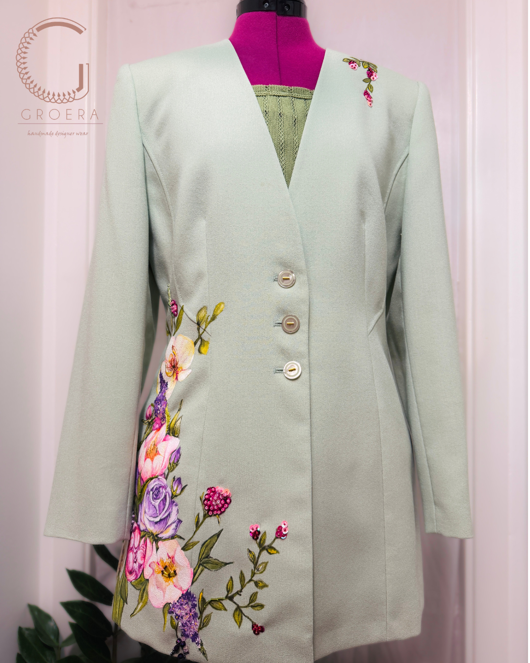 A sage green tailored jacket with colorful floral embroidery on the front and shoulder, displayed on a pink mannequin torso. The jacket features three front buttons and a V-neckline.