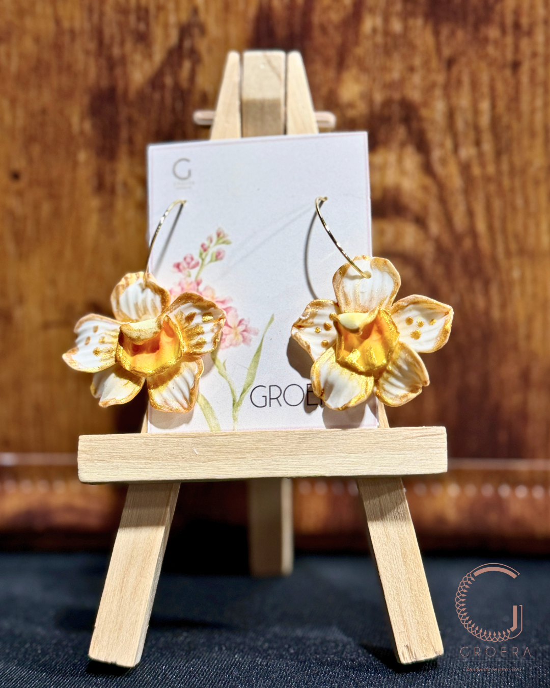 Gold flower-shaped earrings on a small wooden easel with a blurred background