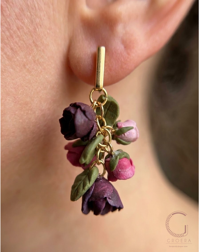A close-up of a person's ear wearing a gold hoop earring adorned with three dangling dried flower charms in shades of purple, pink, and green, with a Groera brand logo in the bottom right corner.