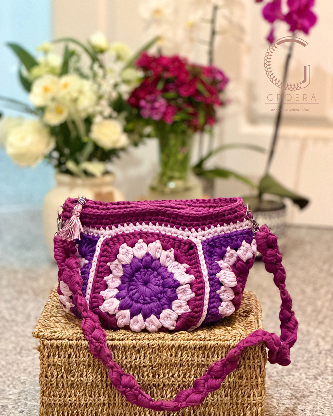 Purple crochet bag with floral patterns on a woven box, flowers in the background
