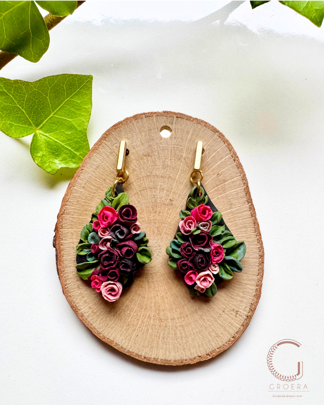 A pair of handmade paper quilling earrings with pink and purple flowers and green leaves, displayed on a round wooden slice stand with green ivy leaves in the background.