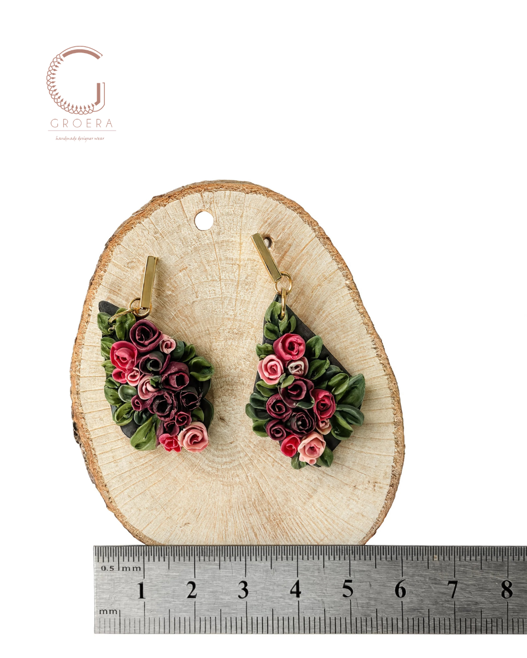 A pair of handmade earrings displayed on a round wood slice, with a metal ruler at the bottom indicating scale. The earrings feature rolled paper flowers in shades of pink, red, and purple with green leaves, hanging from gold-toned posts.