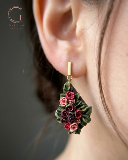 A close-up of a person's ear wearing a dangling flower earring. The earring features sculpted pink and burgundy roses with green leaves on a gold-tone hook, shown against a dark gray background with the Groera brand logo in the upper left.