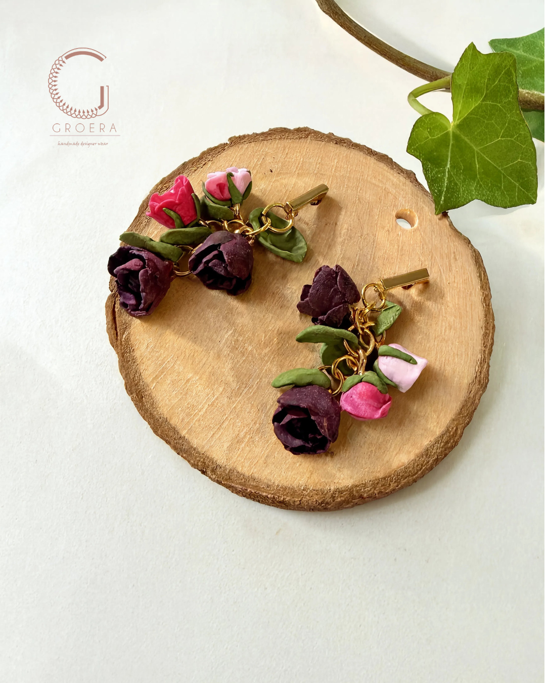 A pair of handmade floral earrings by Groera, featuring dark purple and pink artificial roses with green leaves and gold-tone chains, displayed on a round wood slice.