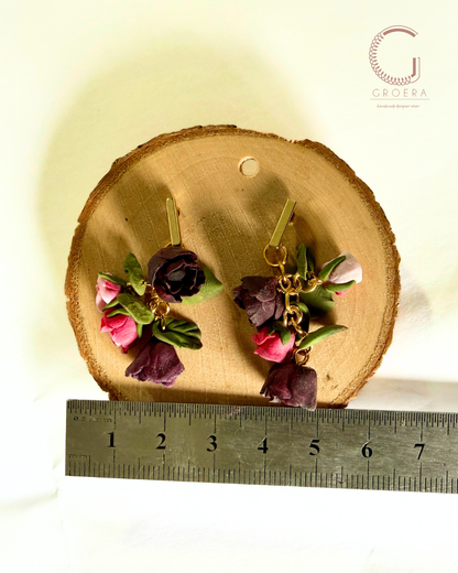 A pair of handmade polymer clay earrings shaped like dark purple roses, pink tulips, and green leaves, hanging from gold-tone hooks. They are displayed on a round wood slice with a metal ruler at the bottom for scale, and a 'GROERA' brand logo in the top right corner.